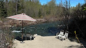 A riverside seating area with a striped umbrella, two white chairs, and a bench.