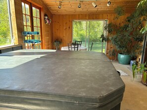 A room with a large bed, wooden walls, a dining table with chairs, and a view of greenery outside.