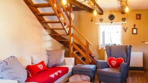 Ein gemütliches Wohnzimmer mit einer Holztreppe, einem grauen Sessel mit Herz-Kissen, einem weißen Sofa mit roten Kissen und einem kleinen Holztisch.