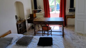 A bedroom with a bed, a desk, and a chair. There is a window with red curtains and a view of greenery outside.