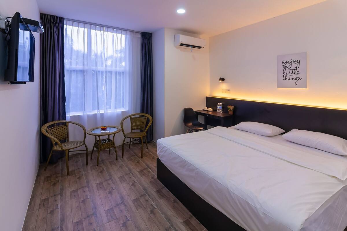 A hotel room with a bed, two wicker chairs, a small table, a desk with a chair, an air conditioning unit, and a wall-mounted TV.
