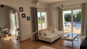 Ein geräumiges Wohnzimmer mit einer Couch, einem Couchtisch und einem Essbereich mit Stühlen. Große Fenster bieten Blick auf einen Pool und Gartenmöbel.