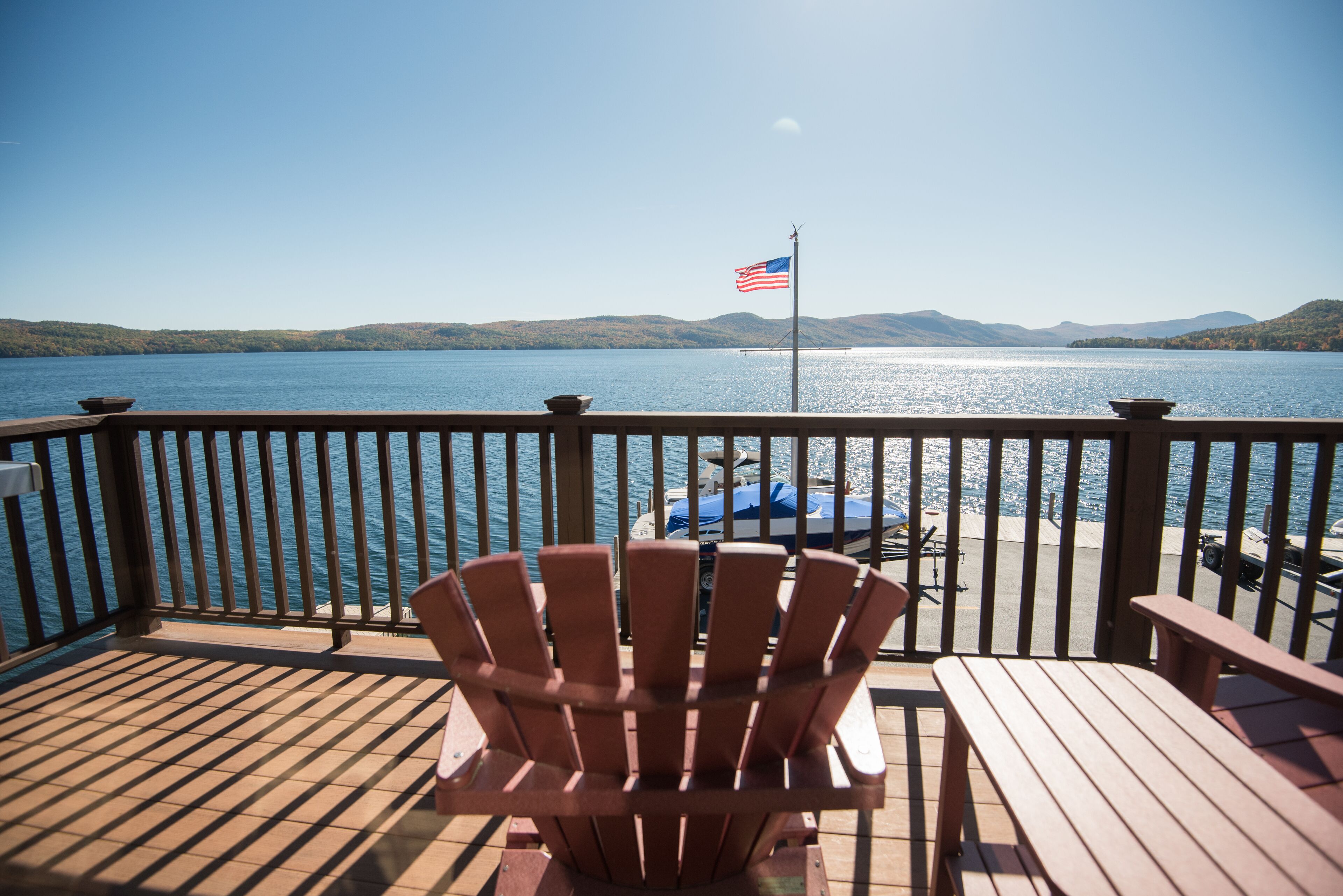 Een houten terras met een rode Adirondack-stoel en een tafel, met uitzicht op een meer met een boot afgemeerd en een Amerikaanse vlag op een paal.