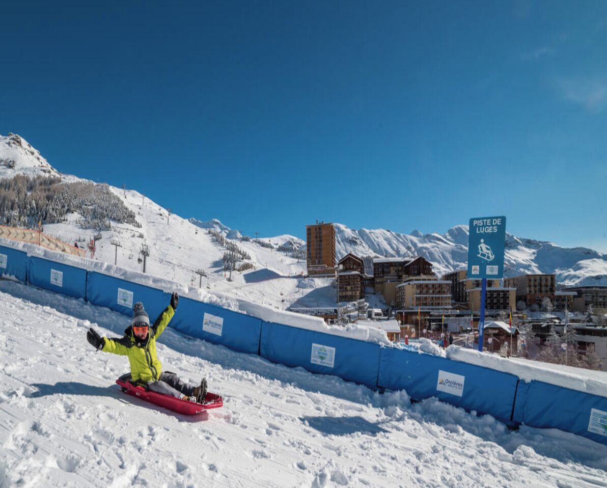 A snow-covered slope with a person snowboarding, a blue barrier, and a building labeled "PISTE DE LUGES" in the background.
