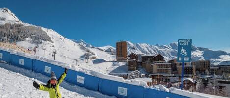 A snow-covered slope with a person snowboarding, a blue barrier, and a building labeled "PISTE DE LUGES" in the background.