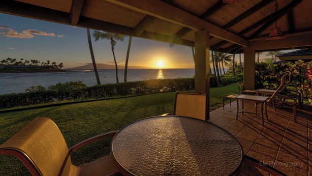 An outdoor patio with a round table and chairs, overlooking a sunset over the ocean with palm trees and a mountain in the distance. - Napili Lani 320 2BD Luxury with Beach Access (Lahaina)