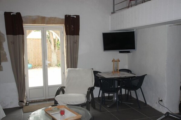 Een kamer met een aan de muur bevestigde televisie, een eettafel met stoelen, een witte fauteuil en een glazen tafel met een dienblad en een rode beker.