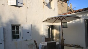 Une terrasse extérieure avec une table et des chaises, un parasol blanc et un bâtiment rustique doté d’un toit en tuiles.
