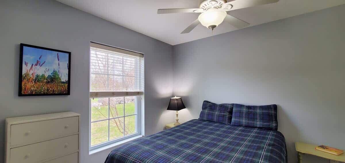 A bedroom with a bed, a dresser, a window with blinds, a ceiling fan, and a painting on the wall.