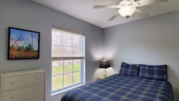 Une chambre à coucher avec un lit, une commode, une fenêtre avec des stores, un ventilateur de plafond et un tableau accroché au mur.