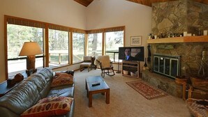 A spacious living room with a stone fireplace, leather sofa, and large windows offering a view of the outdoors. - Air Conditioned/Unobstructed View of Golf Course (Black Butte Ranch)