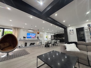A modern living room with a sofa, a coffee table, a dining area with chairs, a kitchen in the background, and a TV mounted on the wall.
