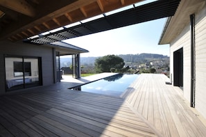 A modern outdoor deck with a wooden floor, a glass-enclosed room, and a view of a hillside town. -  Sarlat 900m from the medieval center, swimming pool, jacuzzi. New ground floor house (Sarlat-La-Canéda)
