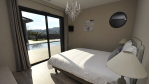 Ein Schlafzimmer mit einem großen Bett, einer Nachttischlampe, einer Kronleuchter, einem runden Spiegel und Blick auf ein Schwimmbad und Grünanlagen durch eine Schiebetür.