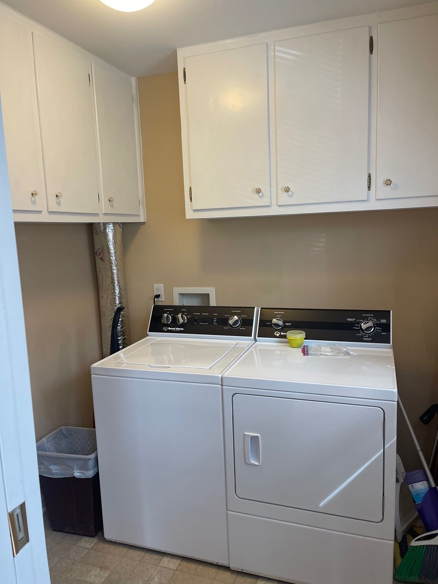 A laundry room with a front-loading washer and dryer, white cabinets above, and a trash bin to the left.