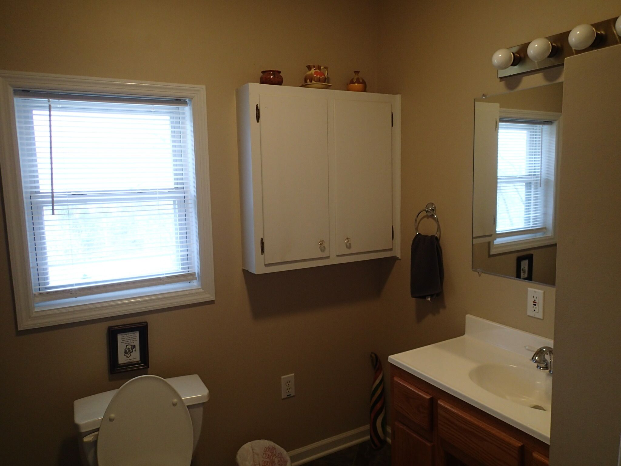 A bathroom with a toilet, sink, mirror, cabinet, and window.