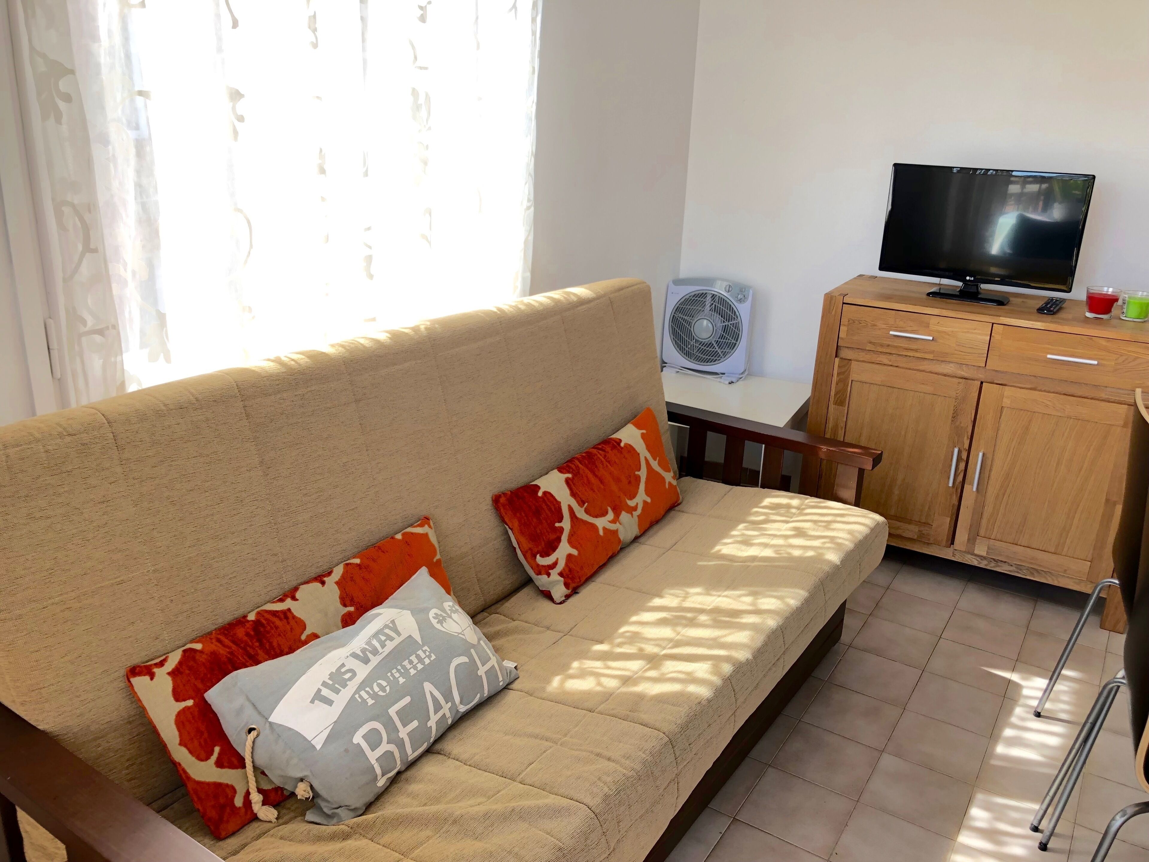 A living room with a beige sofa, two patterned pillows, a television, a wooden cabinet, and a standing fan.