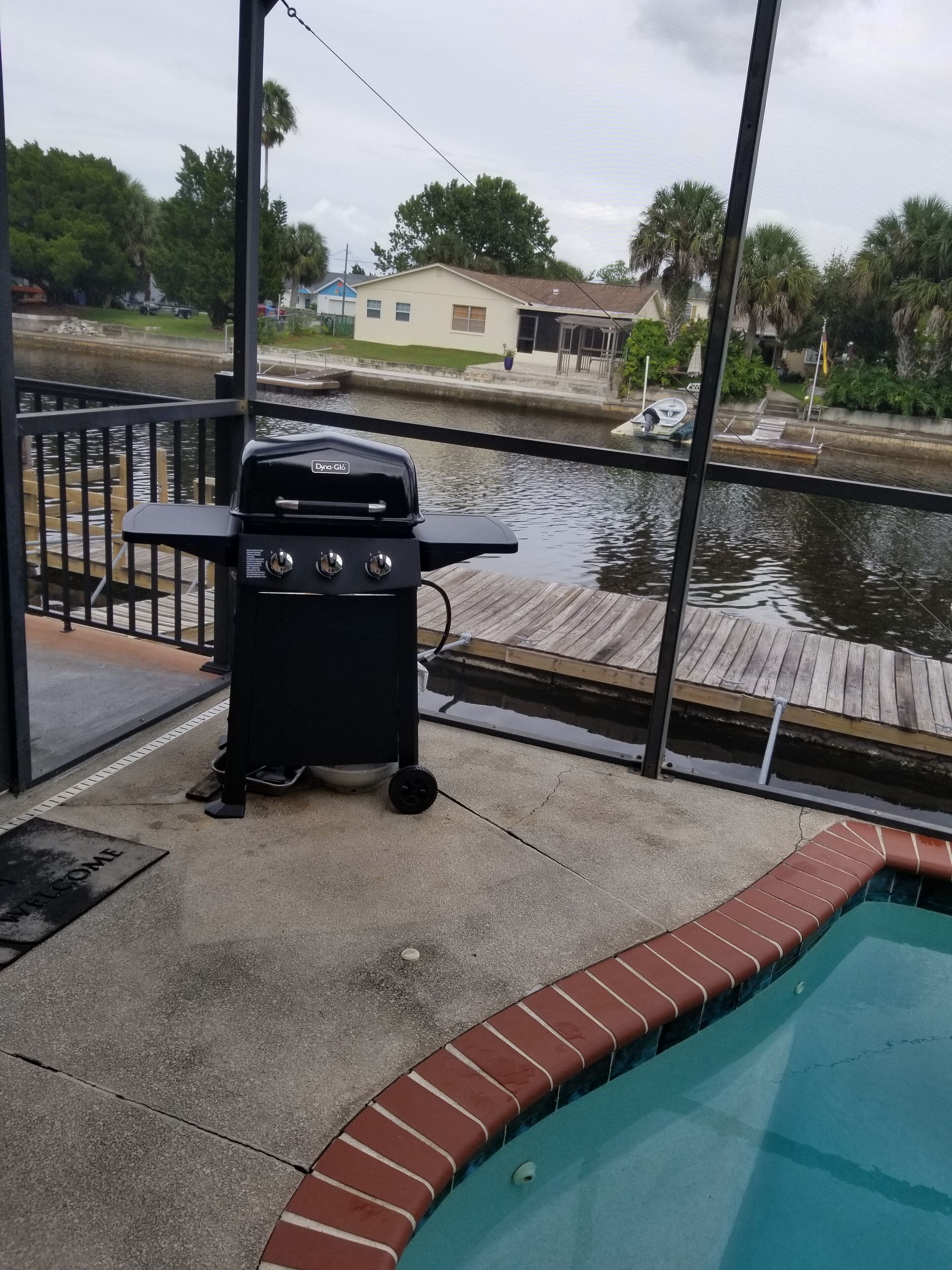A poolside area with a grill, a dock, and a view of a house across the water.