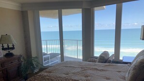 A bedroom with a large bed, a nightstand, a lamp, and a view of the ocean through large windows. - Corner Penthouse - 1101 - Direct Ocean Front with views of Ocean and Intercostal (Daytona Beach)