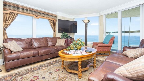 A living room with a brown leather sofa, a wooden coffee table, and a red armchair. A flat-screen TV is mounted on the wall, and there is a large window with a view of the ocean. - Corner Penthouse - 1101 - Direct Ocean Front with views of Ocean and Intercostal (Daytona Beach)