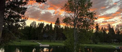 A serene lakeside view with a sunset, trees, and a dock with chairs.