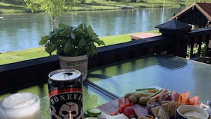 Una mesa con una lata de cerveza, una planta en maceta y una tabla de embutidos, con vista al lago.