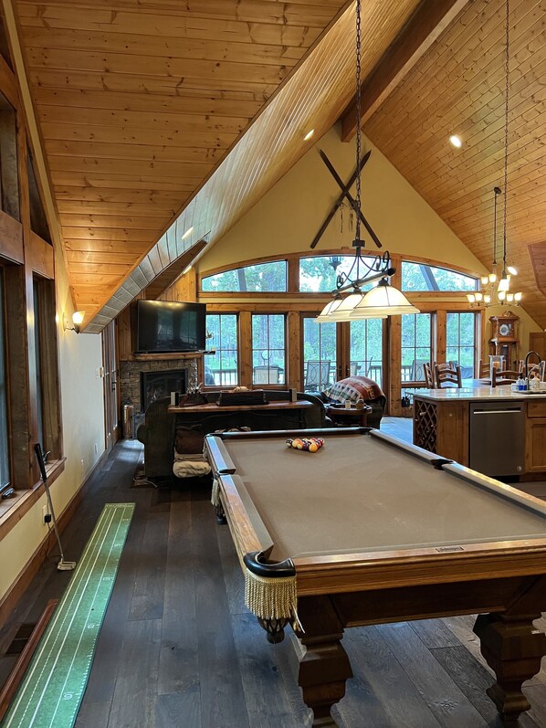 A cozy room with a pool table, wooden ceiling, large windows, and a fireplace.