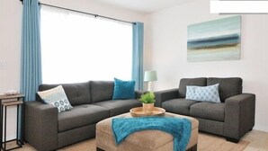 A living room with two grey sofas, a beige ottoman, a small side table, and a window with blue curtains. - New Carlsbad Beach house - Blocks from beach with rare  cental Air Conditioning (Carlsbad)