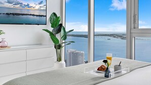 A modern room with a large window overlooking a body of water, a flat-screen TV mounted on the wall, a potted plant, and a breakfast tray with croissant and orange juice.