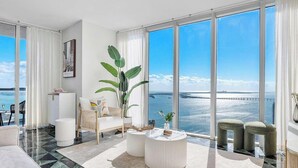 A modern living room with a large window overlooking a body of water and a pier, furnished with a sofa, a coffee table, and a potted plant.