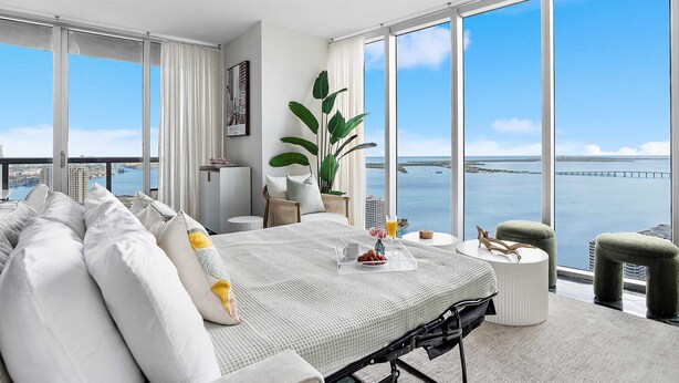 A modern hotel room with a large bed, a dining table, and a view of the water.