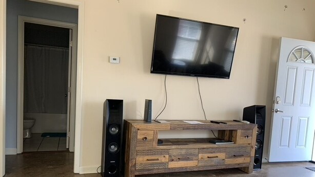 A living room with a wooden entertainment unit, a flat-screen TV, and a door to the right. - Discover Oxford Charm, Just A Quick 5-min Drive To University of Mississippi
 (Oxford)