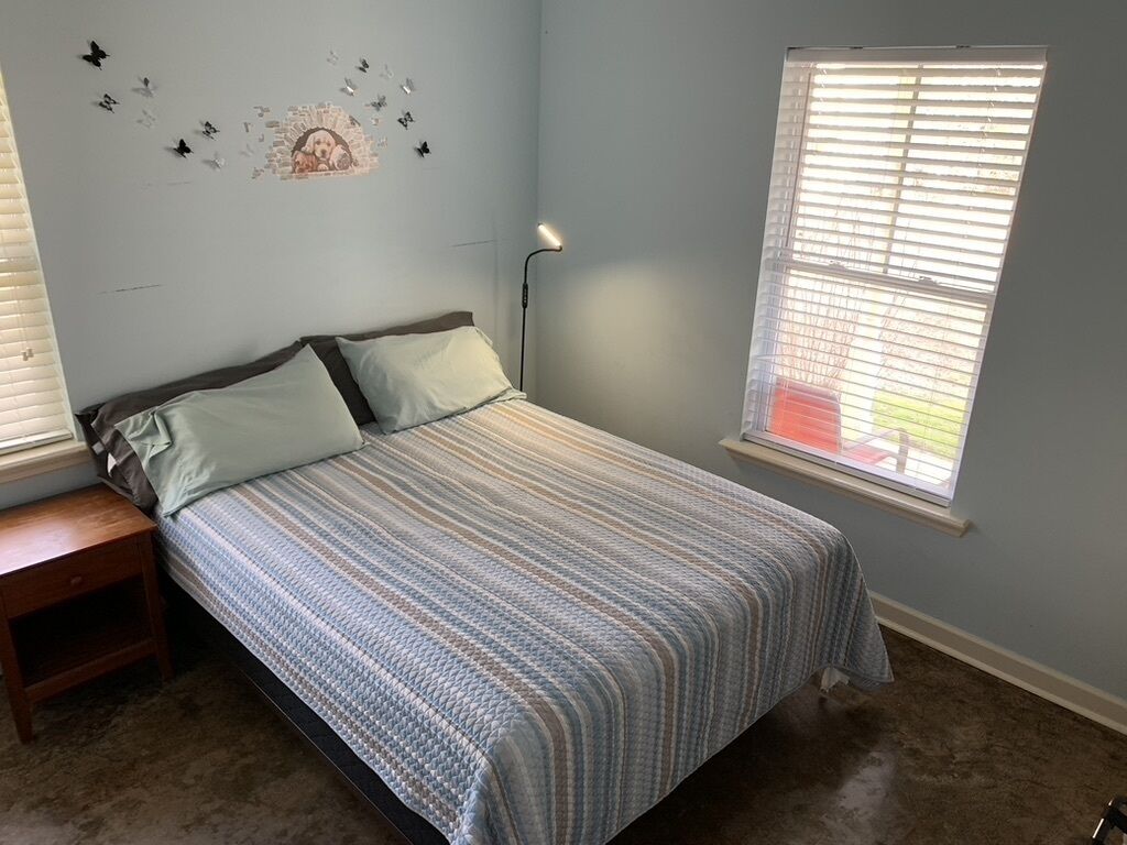 Un dormitorio con cama, mesita de noche, decoración en la pared, ventana con persianas y lámpara.