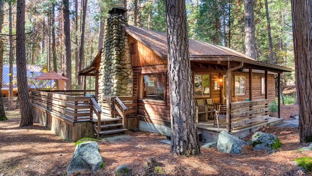 A rustic cabin with a stone chimney, wooden deck, and a porch with seating. - (53) Creekside Cabin at The Redwoods In Yosemite (Yosemite National Park)