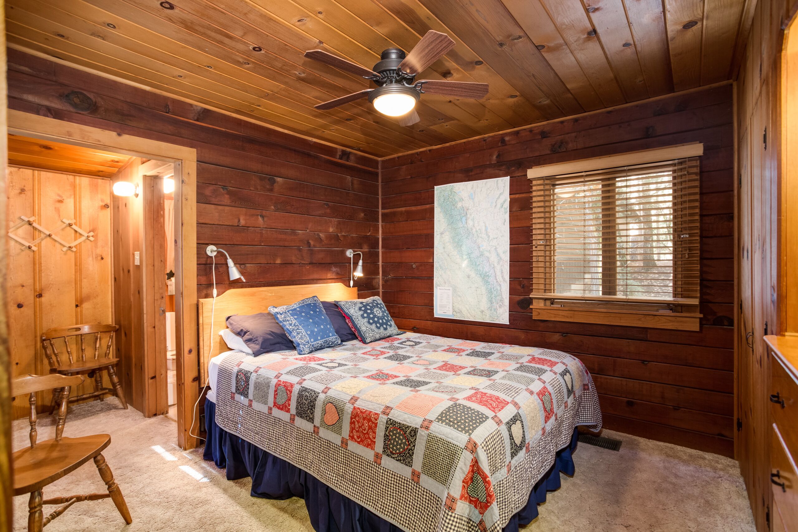 Ein Schlafzimmer einer Holzhütte mit einem Bett, einem Deckenventilator, einem Fenster mit Jalousien und einem Holzstuhl.