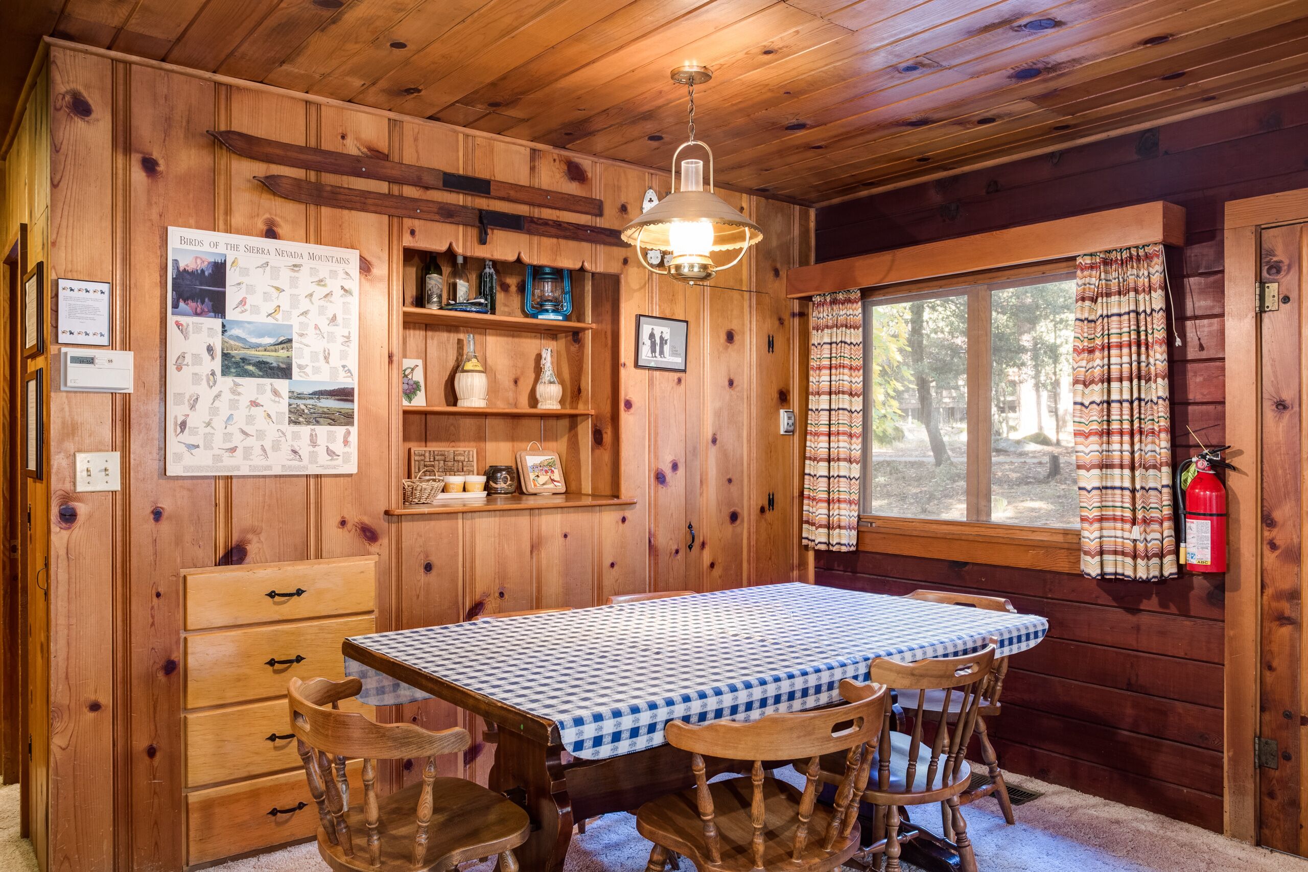 A wooden cabin interior with a dining table, chairs, a hanging light fixture, a window with curtains, and a fire extinguisher.