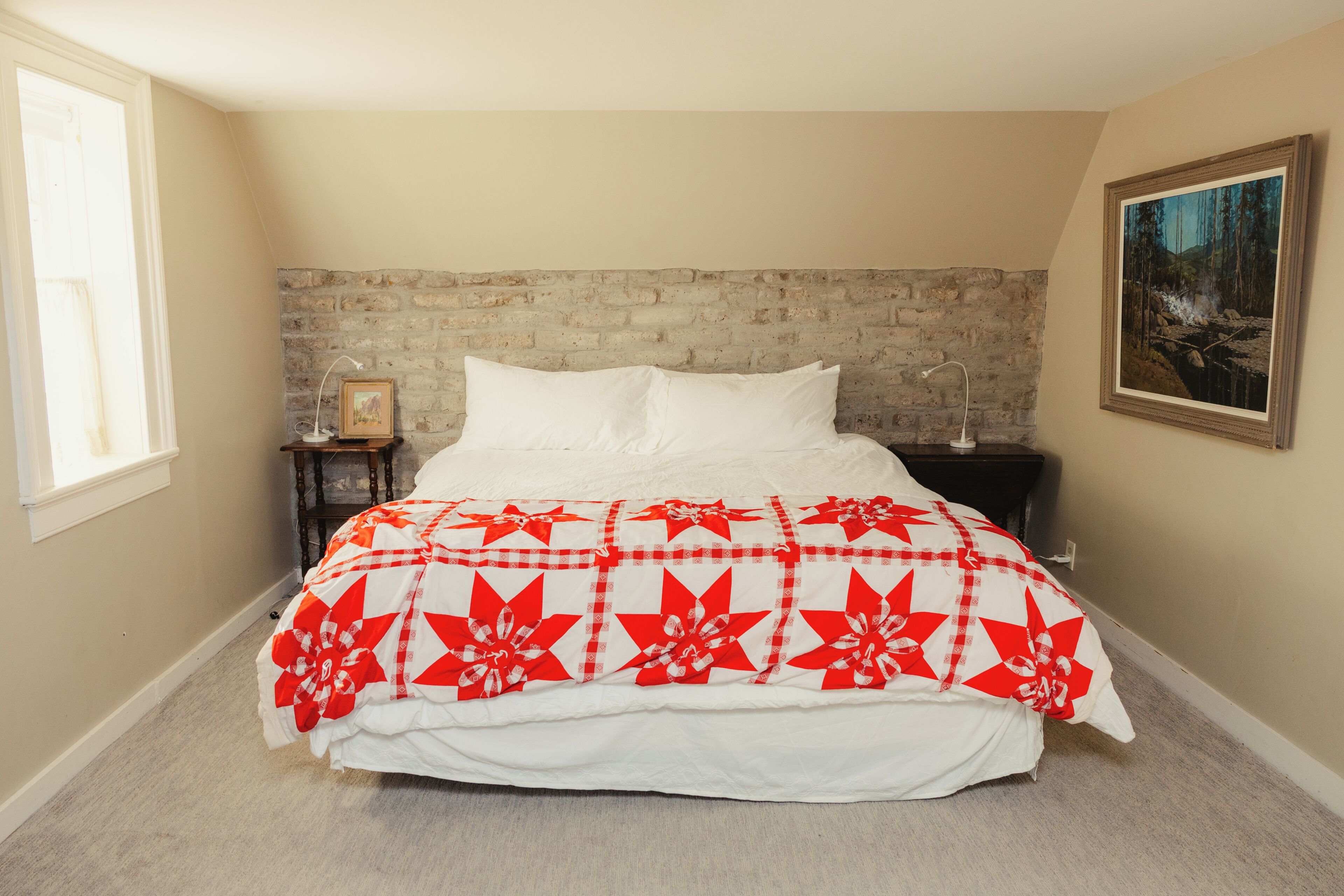Une chambre à coucher avec une tête de lit en briques, un lit recouvert d’une courtepointe à motifs floraux rouges et blancs, une table de chevet avec une lampe et un tableau encadré accroché au mur.