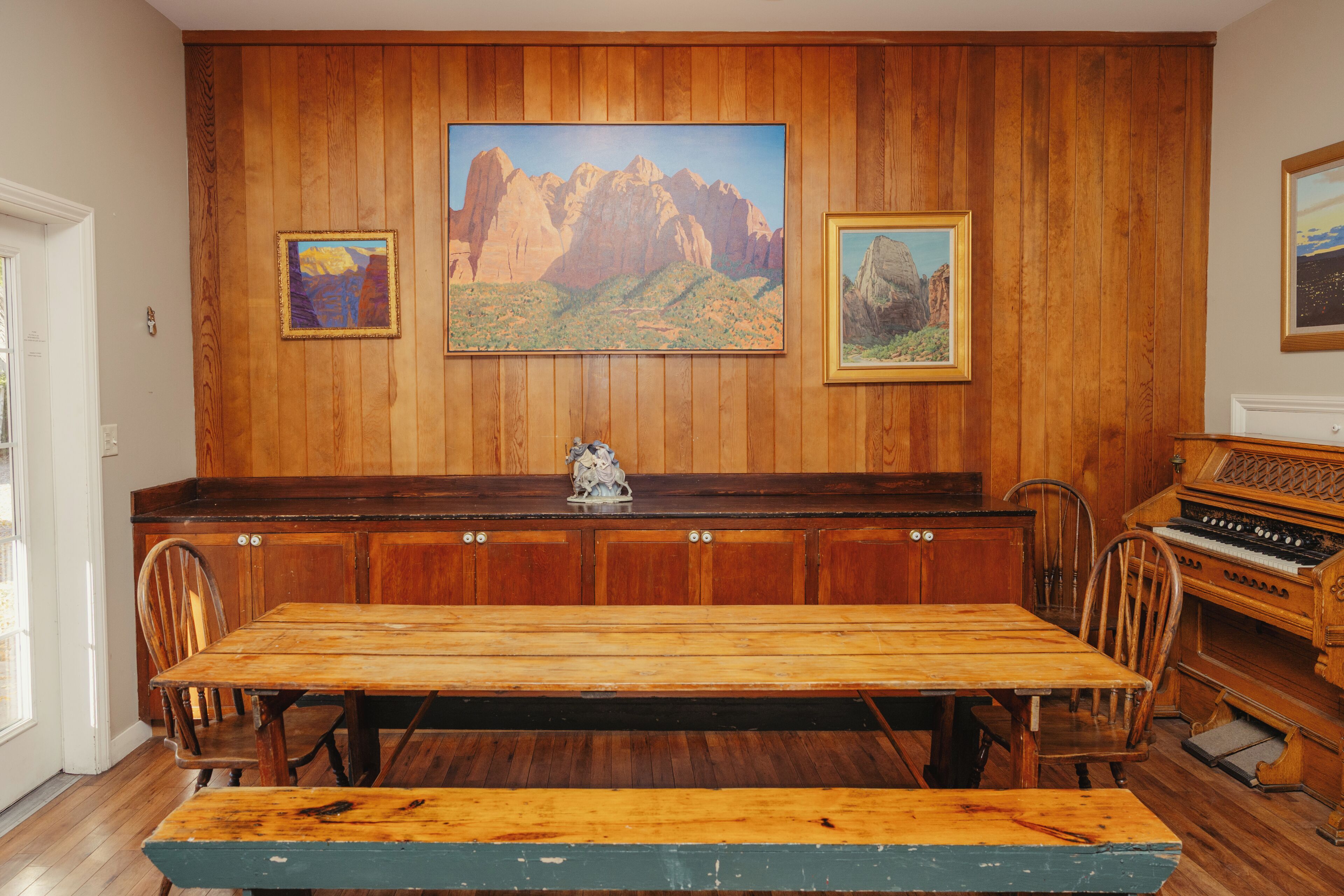 A wooden-paneled room with a dining table, chairs, a piano, and framed paintings on the wall.