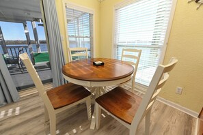A round wooden table with four chairs, a window with blinds, and a view of the water outside.