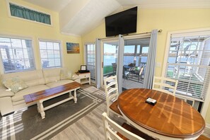 A living room with a sofa, coffee table, and dining table, a TV mounted on the wall, and a view of the water through the windows.