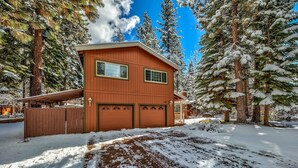 A two-story wooden house with a garage, surrounded by snow-covered trees.