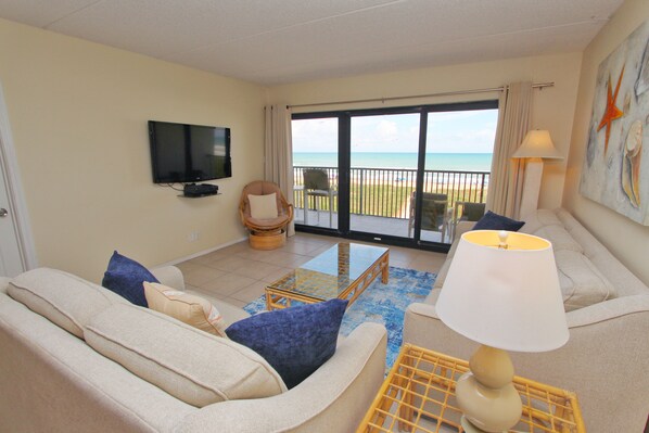 A living room with a sofa, armchairs, a television, and a view of the ocean through a glass door.