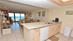 A kitchen with a dishwasher, a sink, and a coffee maker. There is a dining area with chairs and a table. The room has a view of the ocean. - Coastal Fantasea - Condo 403: A spacious 2BR retreat in South Padre Island, perfect for 6! (South Padre Island)