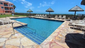 A swimming pool with lounge chairs and umbrellas, surrounded by a stone deck and a building with a red facade. - Coastal Fantasea - Condo 403: A spacious 2BR retreat in South Padre Island, perfect for 6! (South Padre Island)