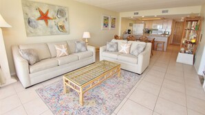 A living room with two sofas, a coffee table, and a kitchen in the background. - Coastal Fantasea - Condo 403: A spacious 2BR retreat in South Padre Island, perfect for 6! (South Padre Island)