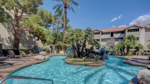 A swimming pool with a waterfall feature, surrounded by lounge chairs and palm trees. - Fantastic Strip view walking distance 2br2b condo (Las Vegas)