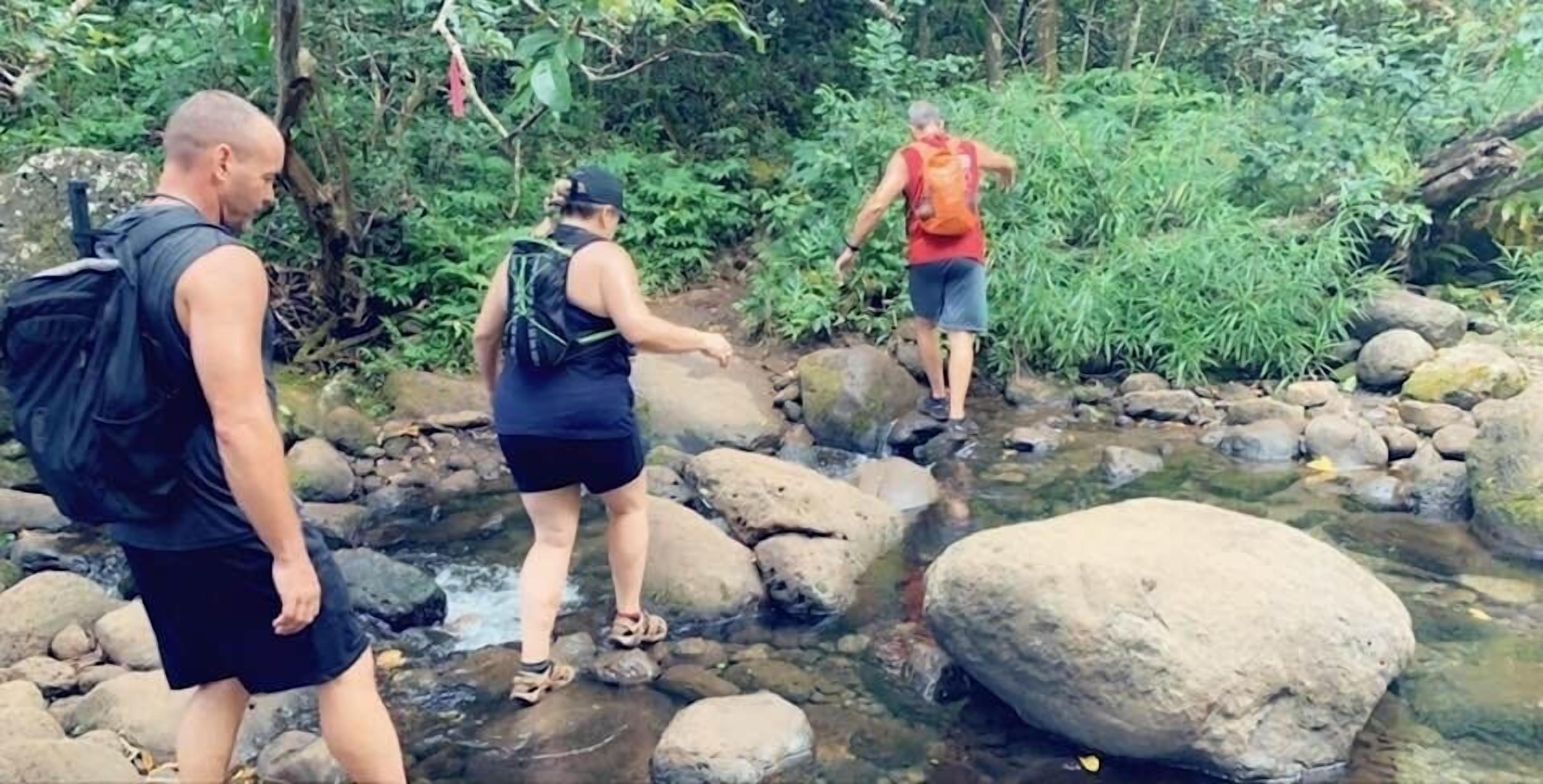 Three people crossing a rocky stream in a forested area.