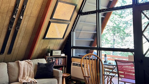 A cozy room with a sloped ceiling, wooden beams, a large window, a sofa, a rocking chair, and a bookshelf.
