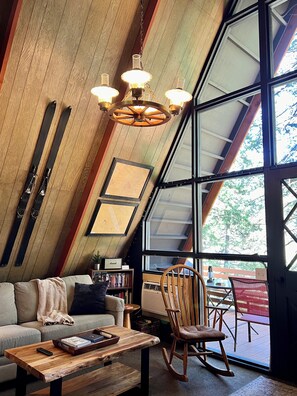 A cozy room with a sloped ceiling, wooden beams, a large window, a sofa, a rocking chair, and a bookshelf.