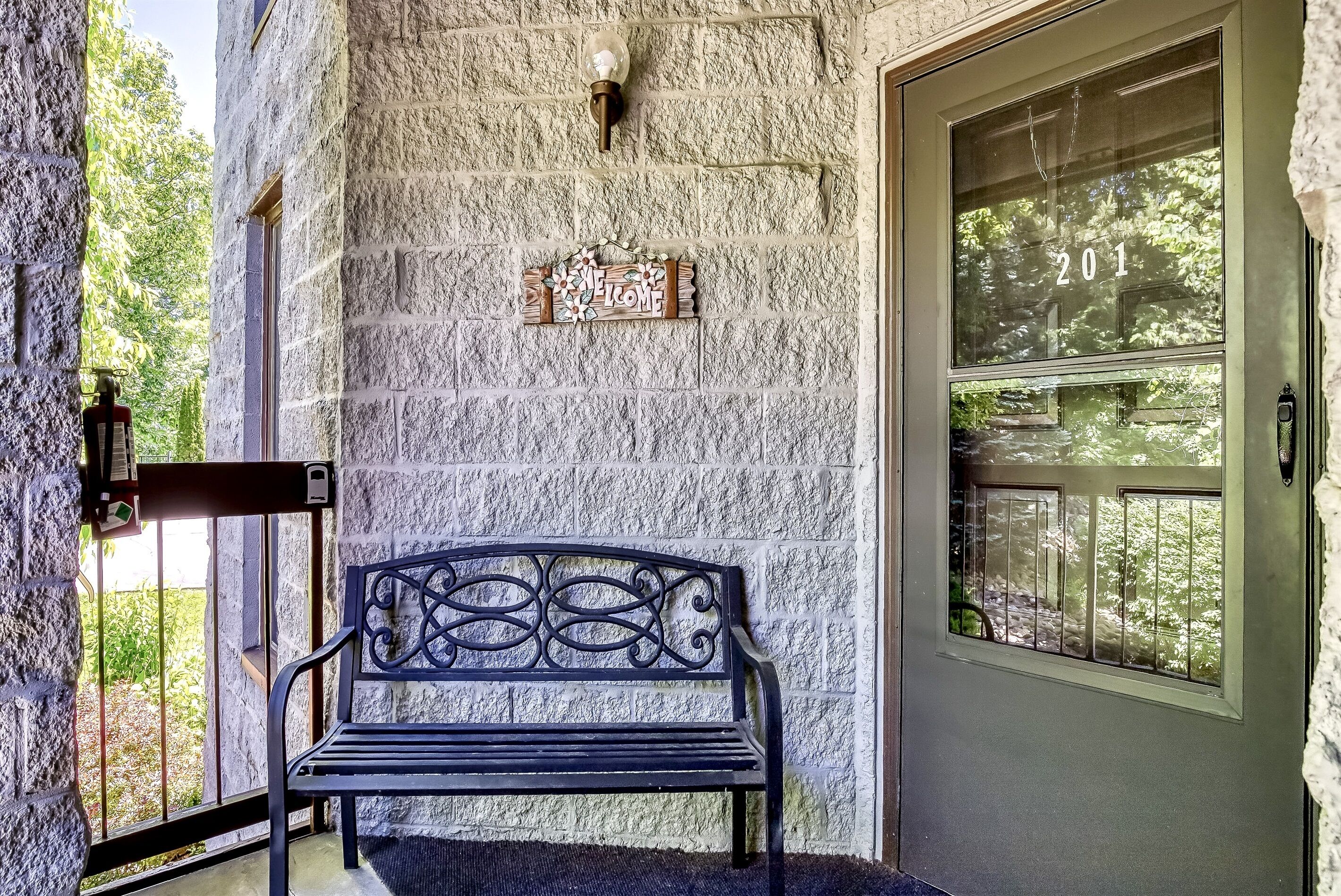 A stone wall with a metal bench, a welcome sign, and a door with a number.